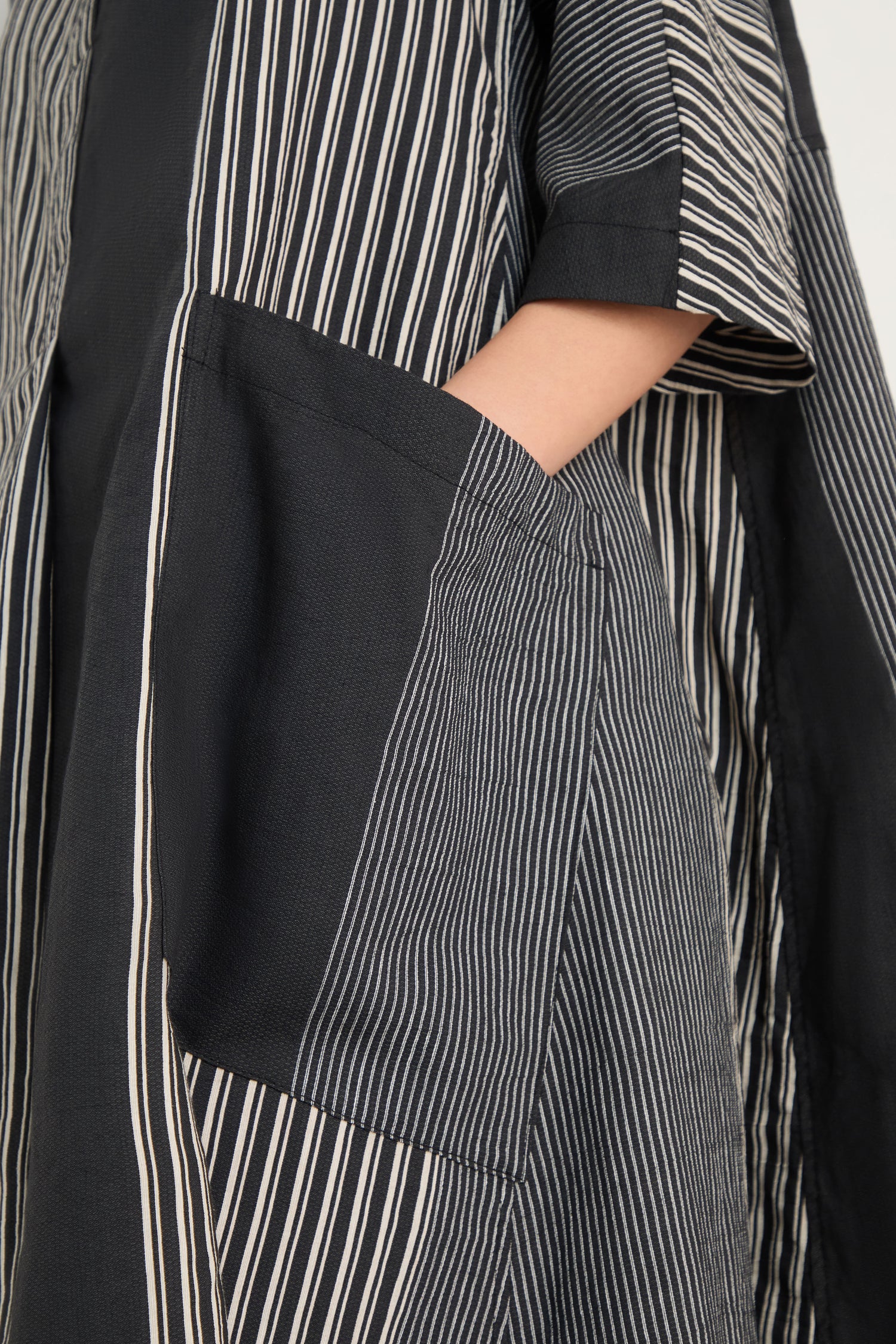 A person’s arm rests by their side while wearing the Mixed Stripe Easy Dress, which features black and white vertical stripes and a large front pocket.