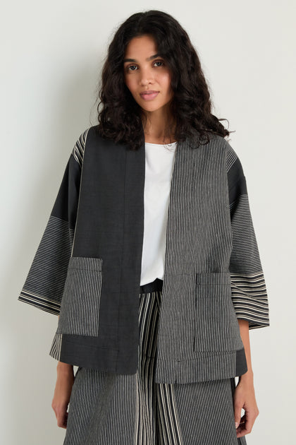 A woman stands against a plain background wearing the Mixed Stripe Easy Jacket in black and gray over a white top with matching striped pants.