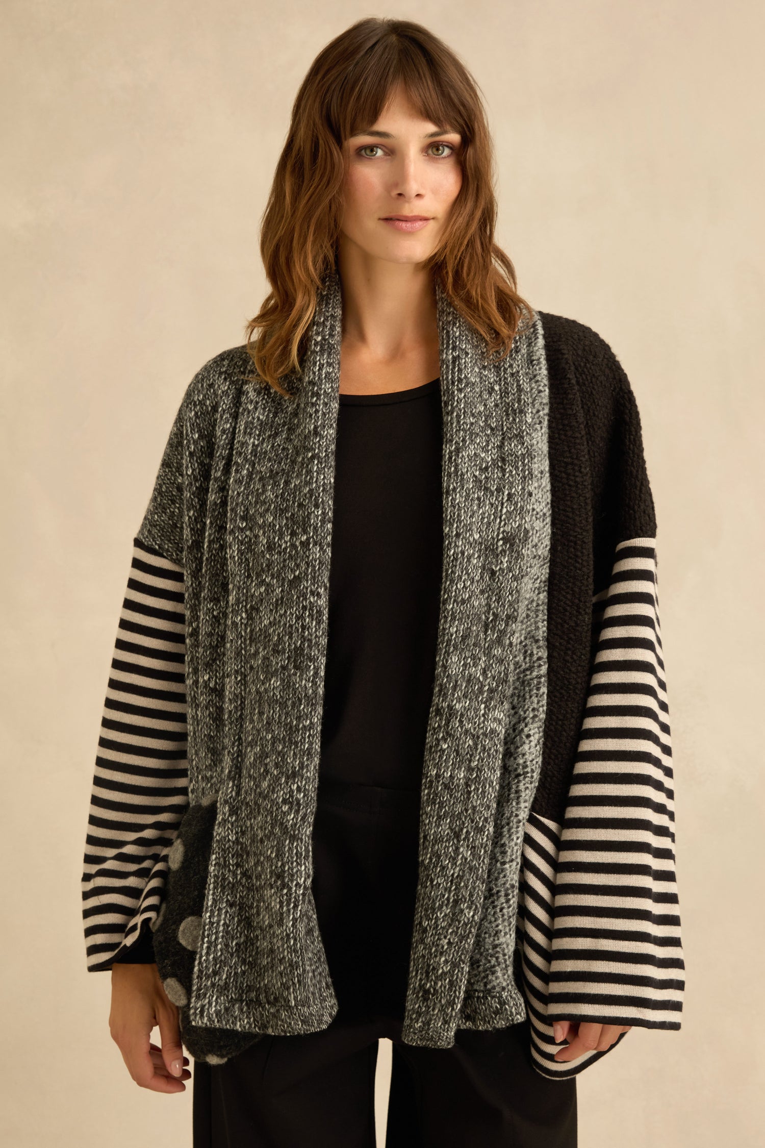 A woman stands against a neutral background wearing the Mixed Spot Stripe Cardigan, its loose fit and combination of gray, black, and striped sections creating visual interest and texture over her black shirt and pants.