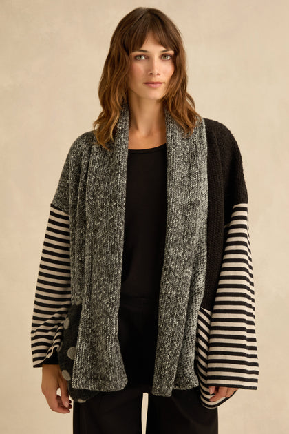 A woman stands against a neutral background wearing the Mixed Spot Stripe Cardigan, its loose fit and combination of gray, black, and striped sections creating visual interest and texture over her black shirt and pants.