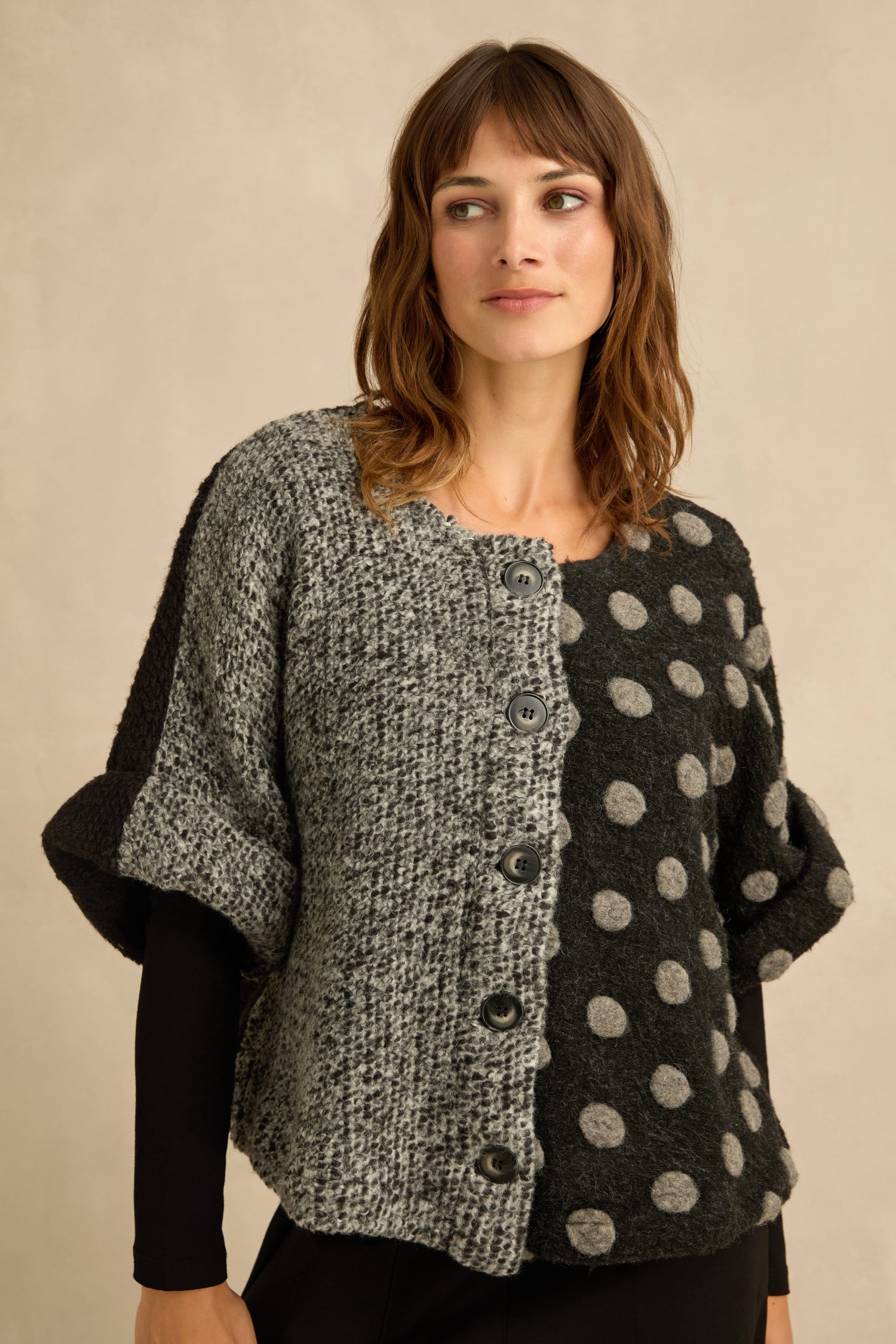 A woman is wearing the Sleeveless Spot Jacket, a mixed textured knit piece with large buttons; the left side displays a tweed pattern while the right side features a polka dot design in gray and black tones.