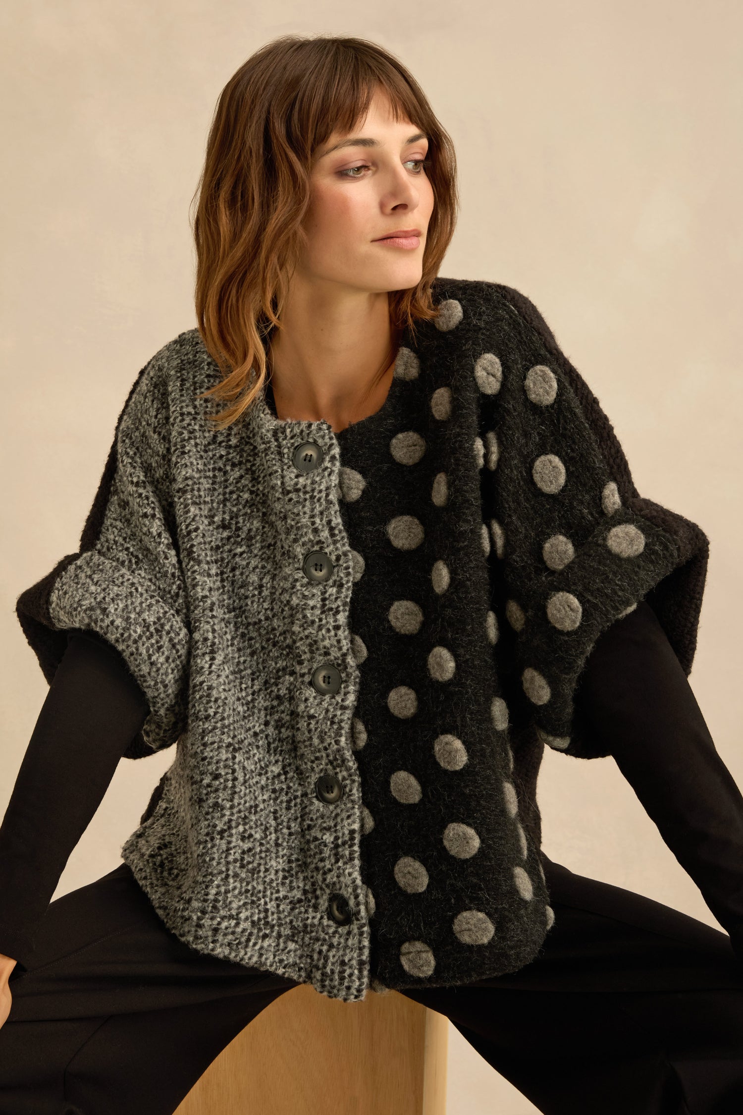 A woman wearing the Sleeveless Spot Jacket—featuring a mixed textured design with one side in gray tweed and the other in black with gray polka dots—sits against a neutral background.