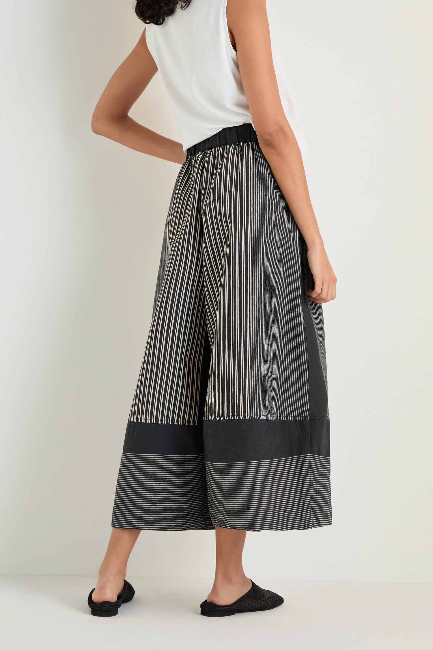 A person stands with their back turned, wearing Mixed Stripe Wide Leg Trousers featuring an elasticated waist, paired with a sleeveless white top and black flats.