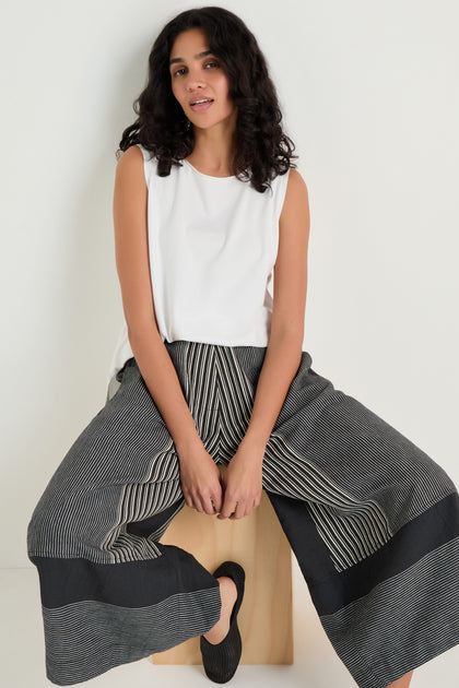 A woman with curly hair sits on a wooden bench in a sleeveless white top, black shoes, and Mixed Stripe Wide Leg Trousers against a plain light background.