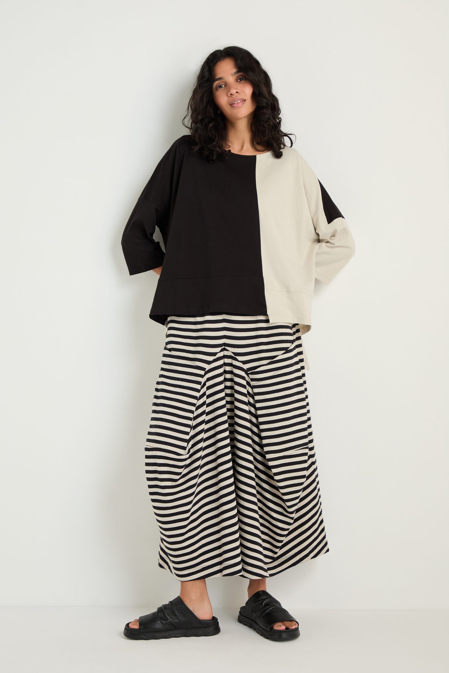 A person stands against a white wall wearing the Two-Tone Jersey Top, wide black-and-white striped pants, and black sandals.