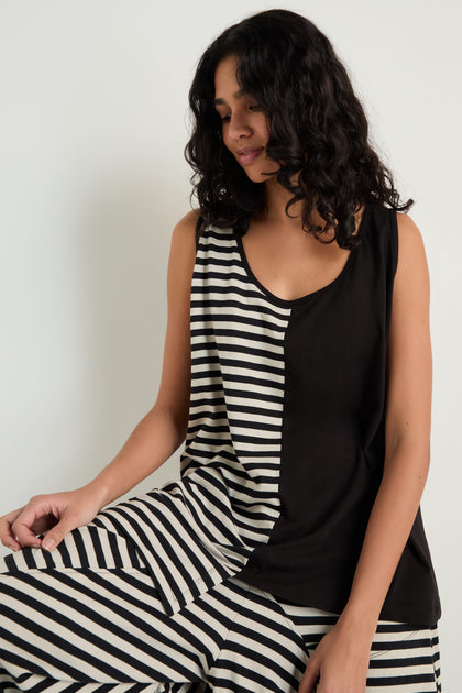 A woman with curly hair wears the Stripe and Plain Sleeveless Top, featuring black and white cotton stripe panels, paired with pants against a plain light background.