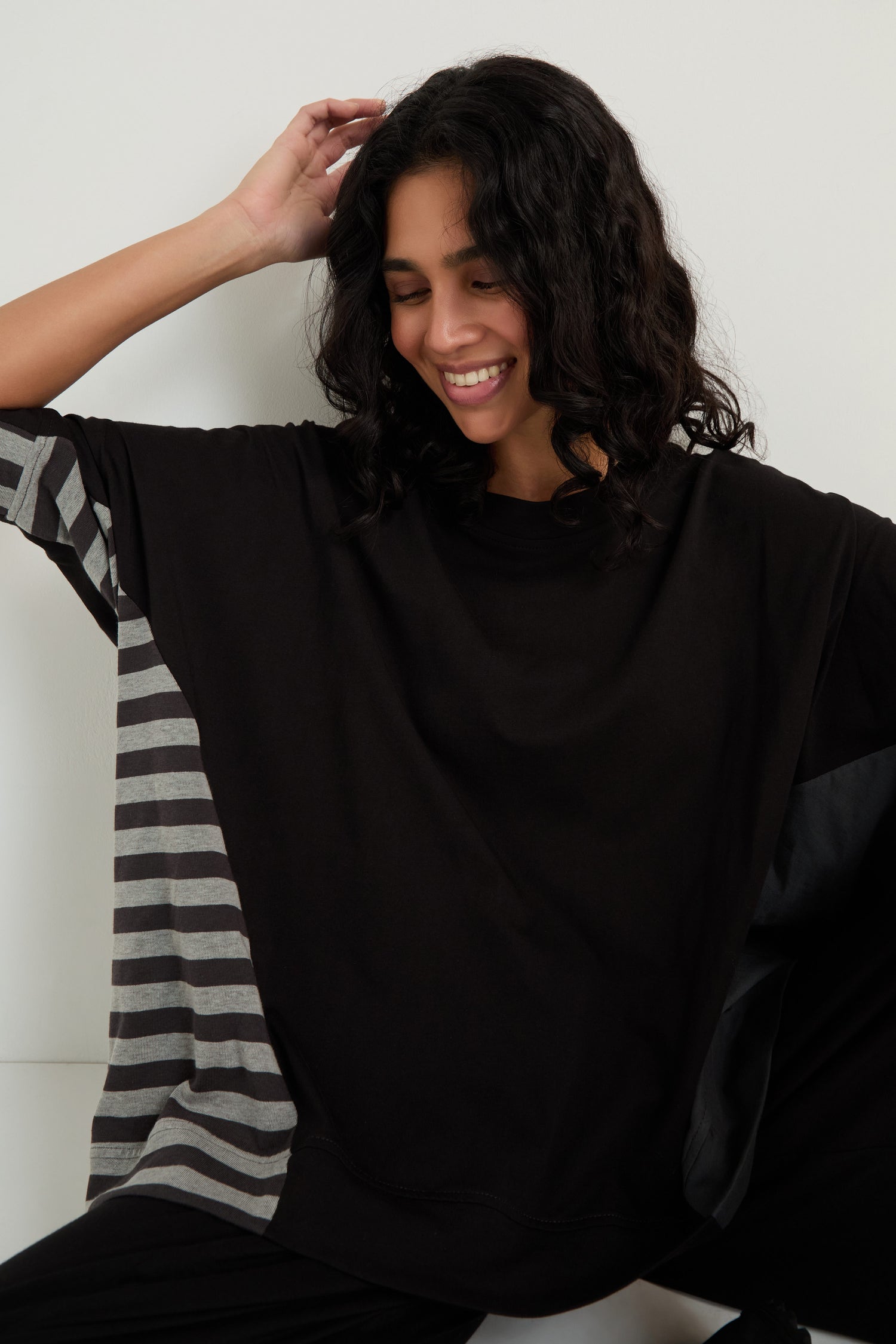 A person with curly dark hair smiles while wearing the Boxy Side Detail Top with gray and black striped sleeves, seated against a white background.