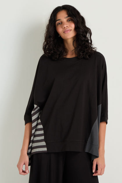 A woman stands against a light-colored wall, wearing the Boxy Side Detail Top with gray striped panel accents and black pants.