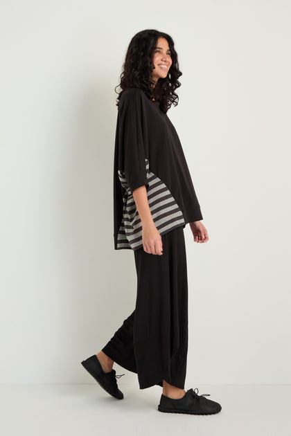 A person stands sideways, smiling, wearing the Boxy Side Detail Top with gray side stripes, wide black pants, and black shoes against a plain white background.