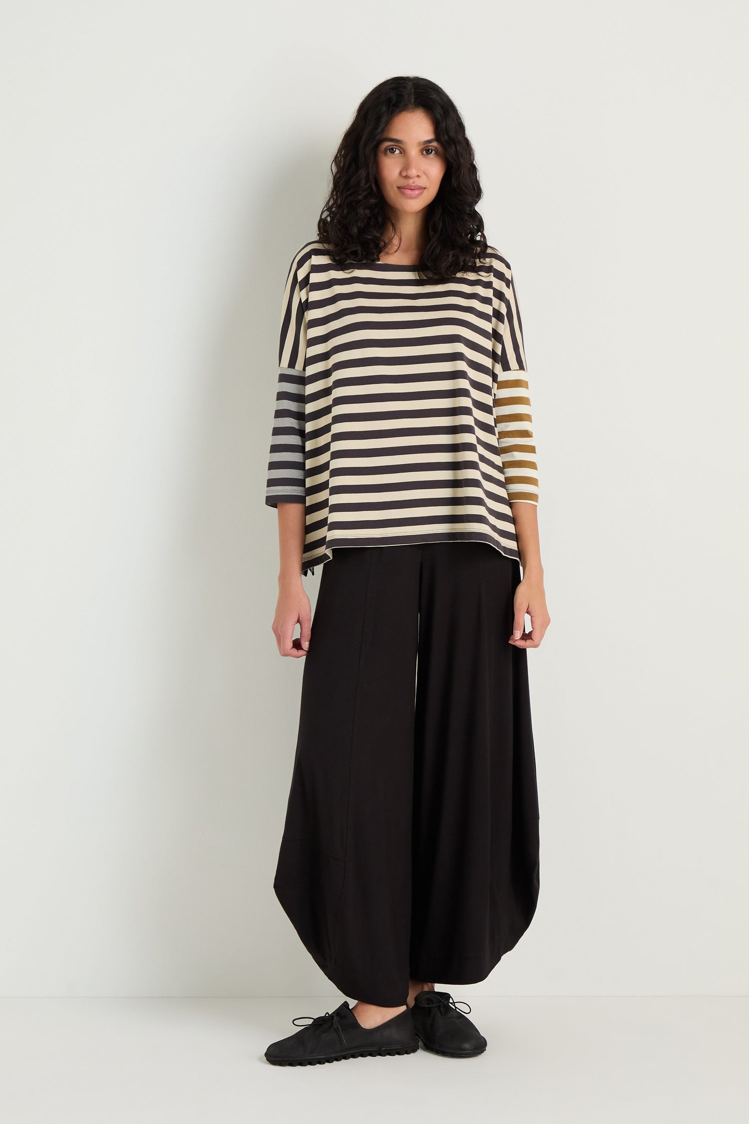 A person stands against a plain wall wearing the Boxy Stripe Top with three-quarter sleeves and wide black pants, creating a relaxed silhouette completed with black shoes.
