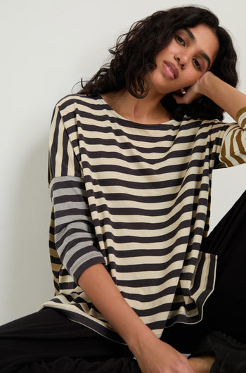 A woman with wavy dark hair rests her head on her hand, wearing the Boxy Stripe Top and black pants, creating a relaxed look against a plain background.