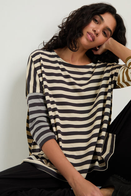 A woman with wavy dark hair rests her head on her hand, wearing the Boxy Stripe Top and black pants, creating a relaxed look against a plain background.