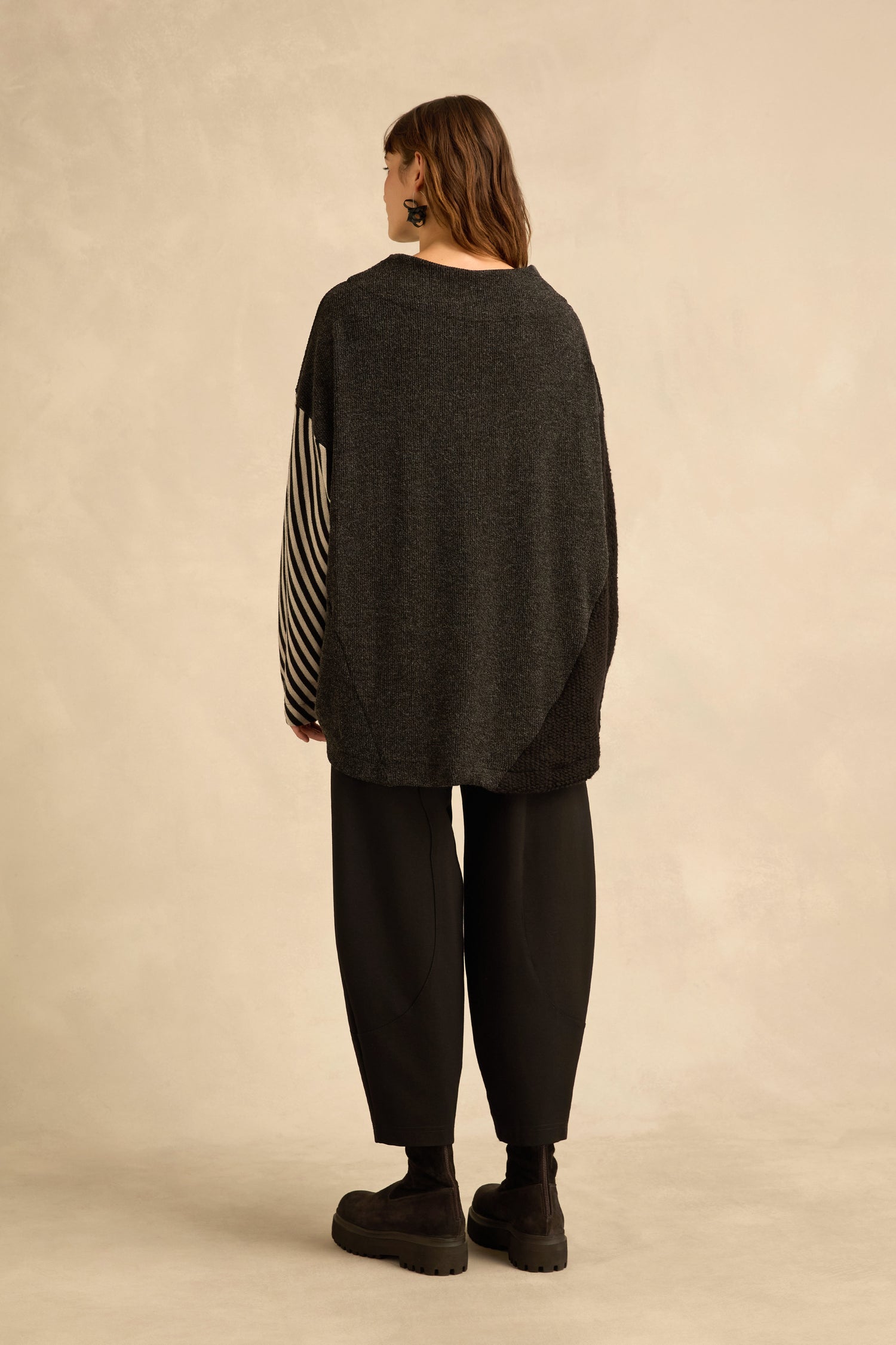 A person stands with their back to the camera, wearing the Mixed Stripe Top—a charcoal oversized sweater with asymmetric panel construction and one striped sleeve—paired with loose black pants and black boots against a neutral background.