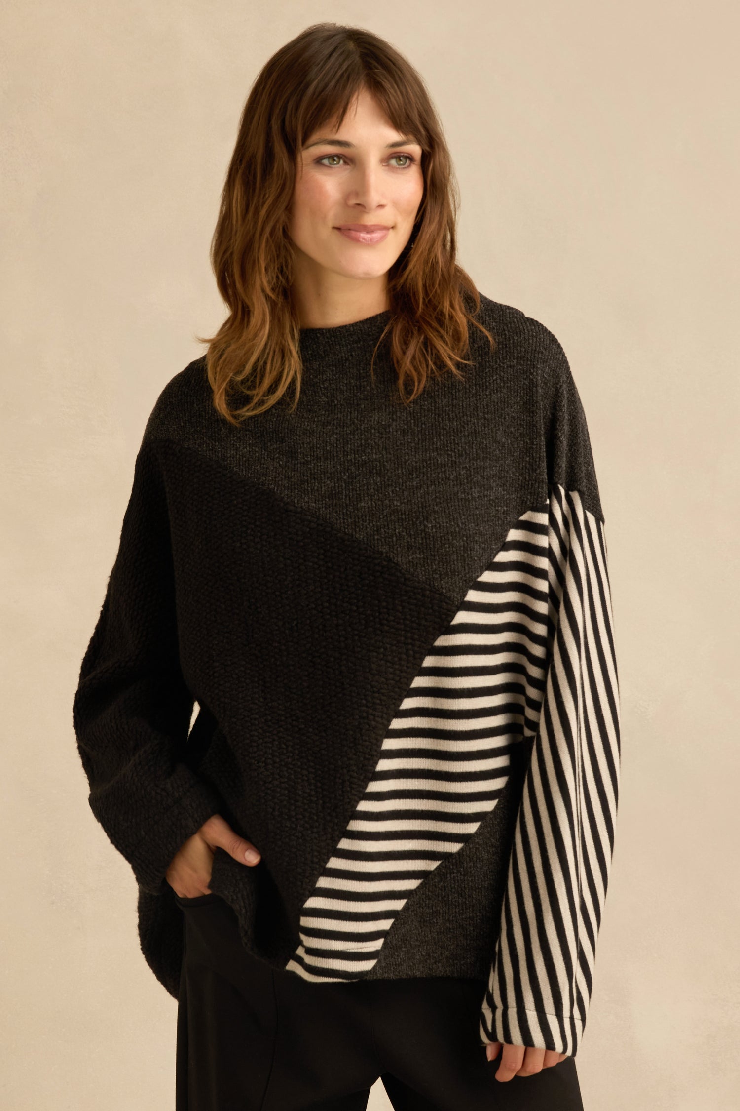 A woman is wearing the Mixed Stripe Top—an oversized black sweater with asymmetric panel construction and diagonal black and white striped panels on one sleeve and part of the front—standing with one hand in her pocket.