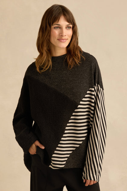 A woman is wearing the Mixed Stripe Top—an oversized black sweater with asymmetric panel construction and diagonal black and white striped panels on one sleeve and part of the front—standing with one hand in her pocket.