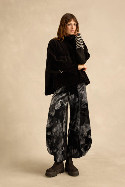 A woman stands against a neutral background, wearing the Cowl Neck Velvet Jersey Top by Urban Alembika, paired with wide-leg black and gray tie-dye pants and black platform boots.