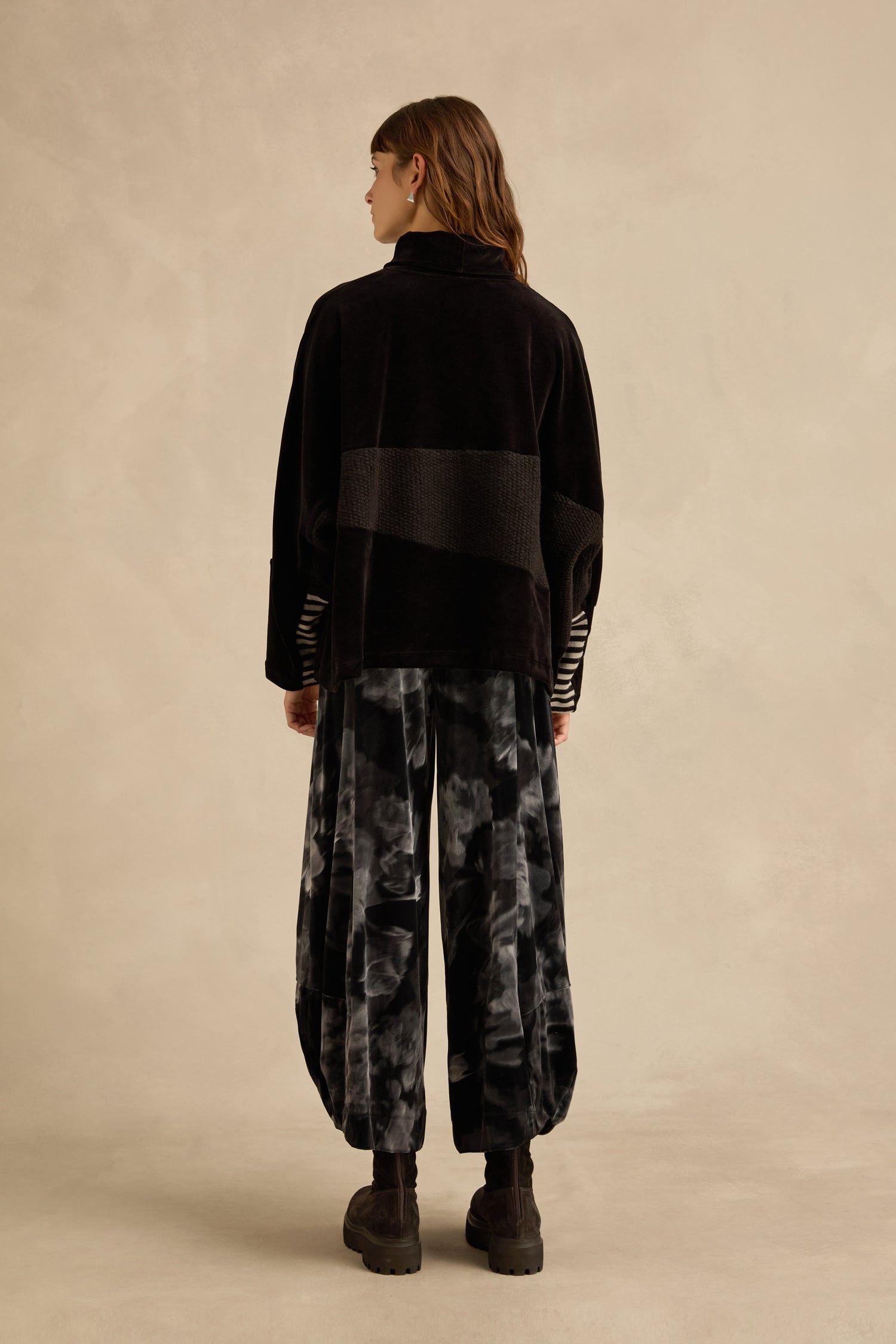 A person stands with their back to the camera, wearing a black Cowl Neck Velvet Jersey Top, loose tie-dye pants, and black boots against a neutral beige background.
