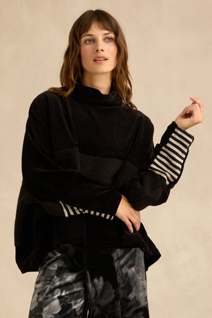 A woman models the Cowl Neck Velvet Jersey Top, featuring a loose black cowl neck and striped sleeves, paired with black-and-white tie-dye pants and set against a neutral background.