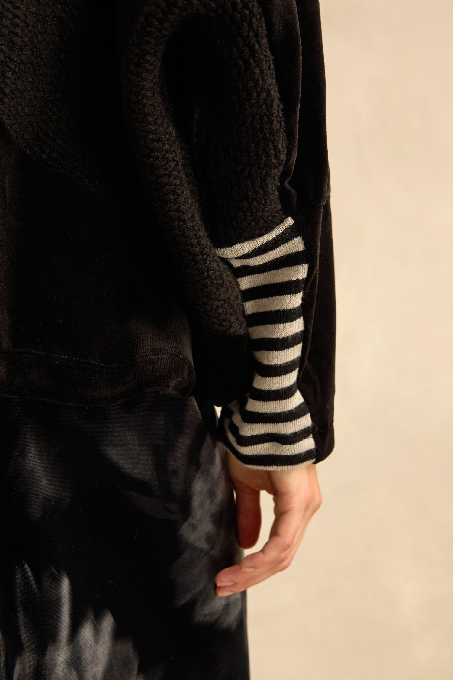 Wearing the Cowl Neck Velvet Jersey Top, a person stands with their arm relaxed, layered underneath a dark patterned skirt and paired with a black textured jacket featuring striped sleeves.