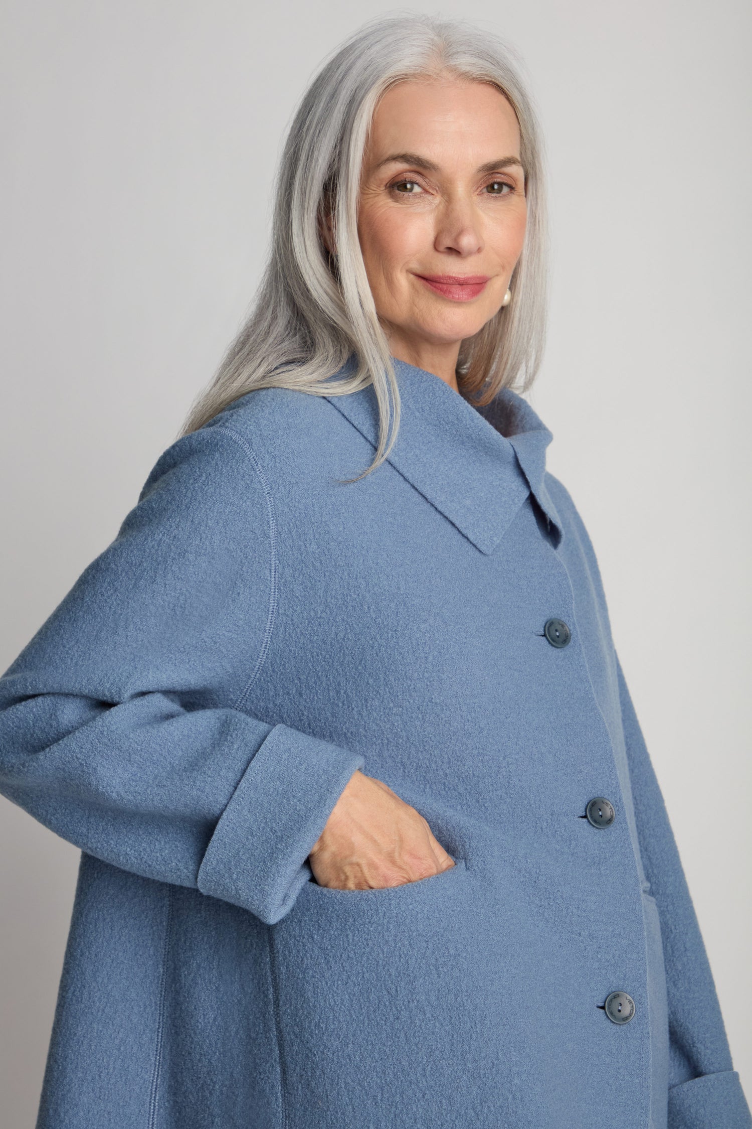 A woman with long gray hair wearing a light blue Collar Boiled Wool Jacket, standing with her hand in the jacket pocket and smiling slightly.