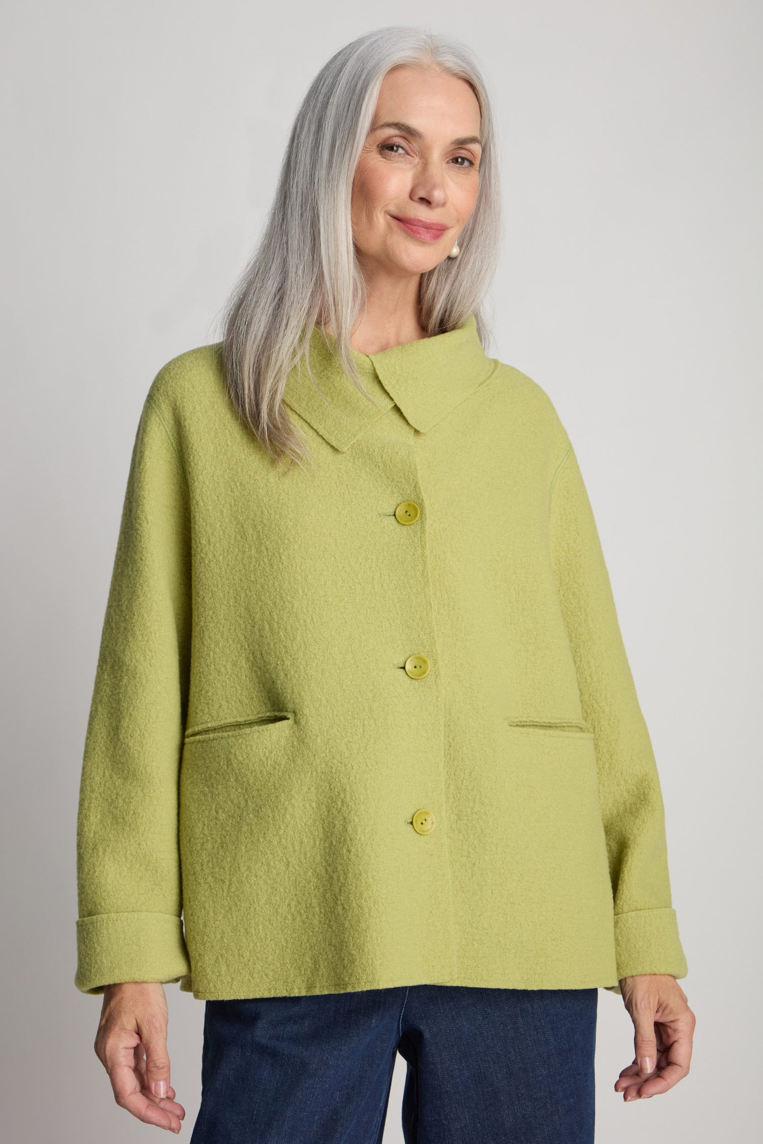 A woman with long grey hair, dressed in a light green Collar Boiled Wool Jacket and blue jeans, stands in front of a plain background.