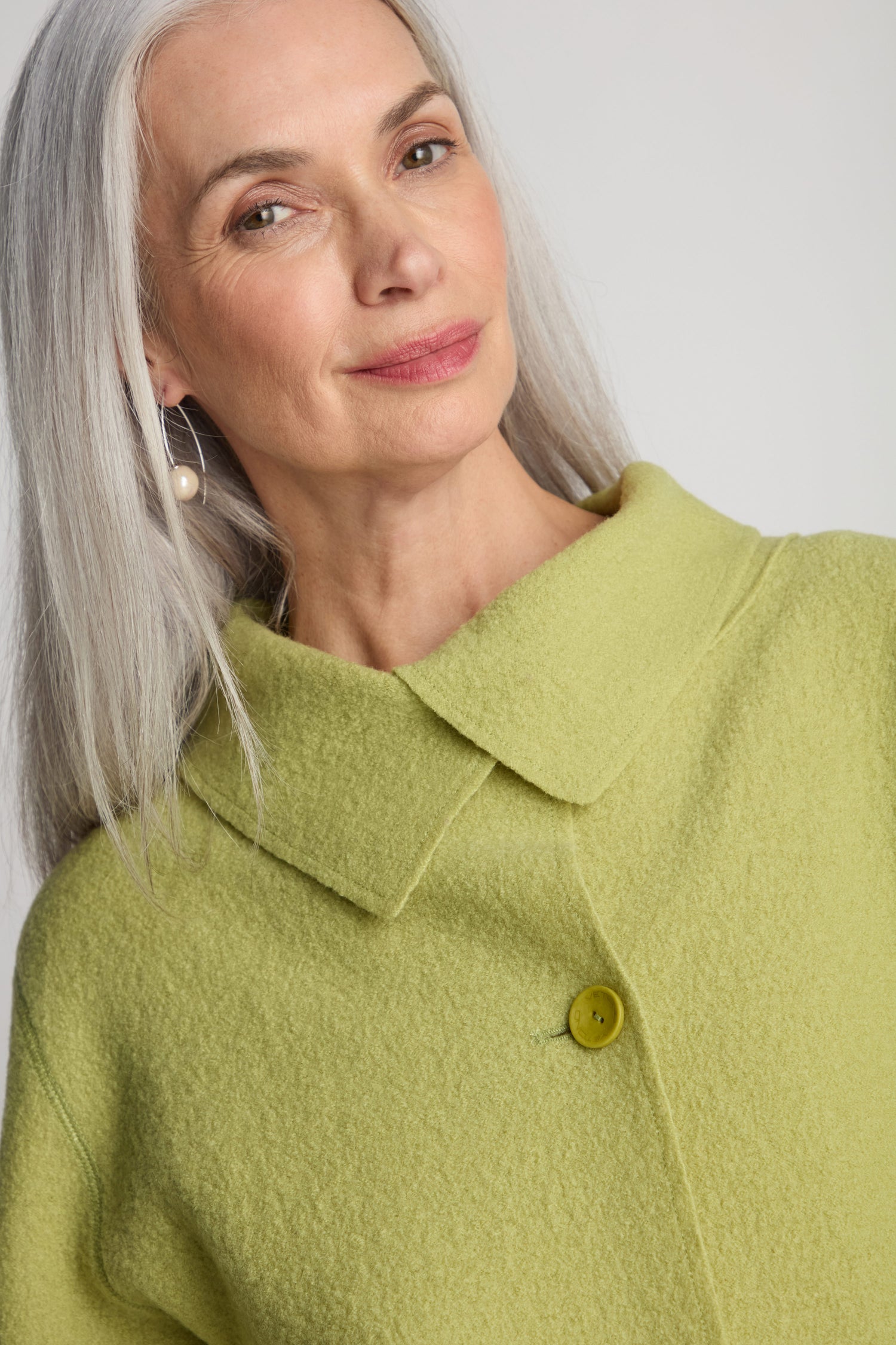 A woman with long gray hair, donned in an oversized Collar Boiled Wool Jacket in yellow-green, stylishly accented with a chic collar and paired with pearl earrings, is smiling slightly while looking into the camera.