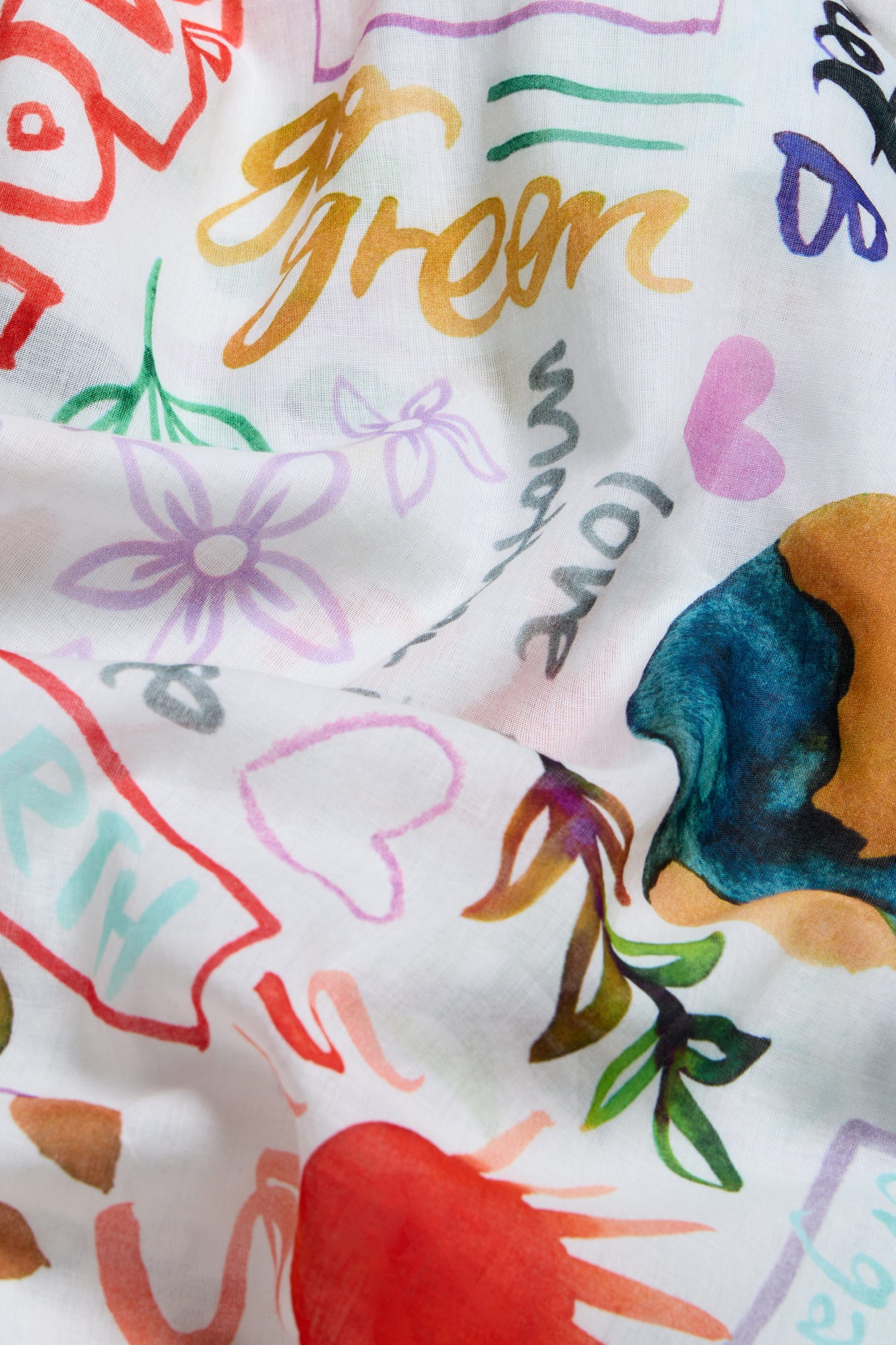 The Peace and Love Cotton Scarf is a lightweight white scarf with vibrant floral prints, colorful doodles, hearts, abstract shapes, and playful words like "green," "love," and "earth.