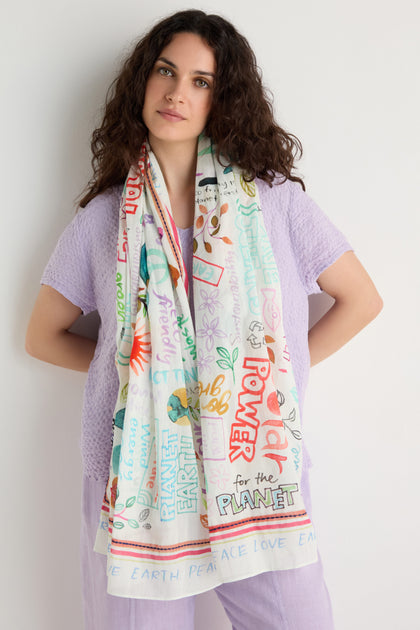A woman with curly hair in a light purple outfit wears the Peace and Love Cotton Scarf, featuring colorful eco-themed words and drawings, standing against a plain white background.