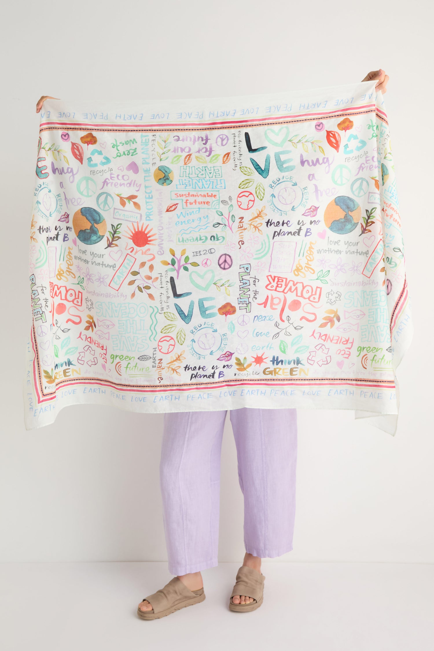 A person displays the Peace and Love Cotton Scarf, featuring vibrant illustrations and words about peace, love, the planet, and nature. They are dressed in lavender pants and beige sandals.
