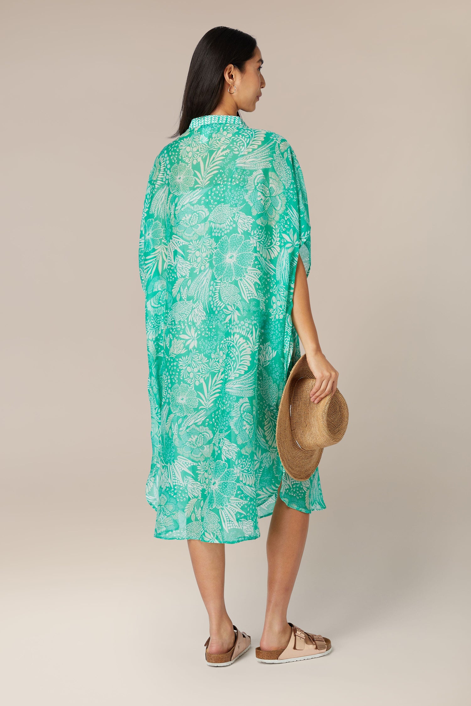 A person standing in profile wearing a green Button Bouquet shirt dress and holding a straw hat.