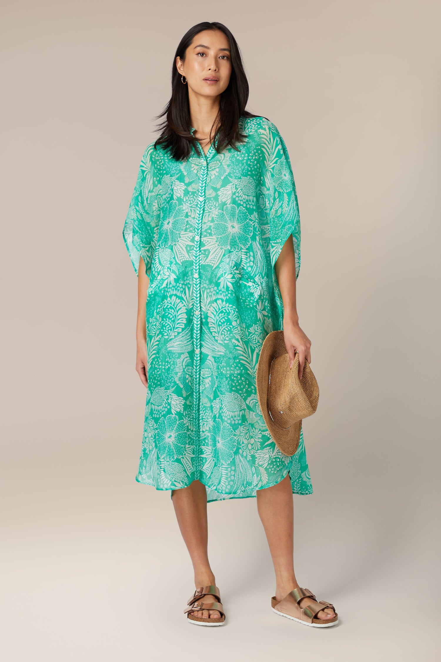 A woman in a green patterned Button Bouquet Shirt Dress holding a hat stands against a neutral background.