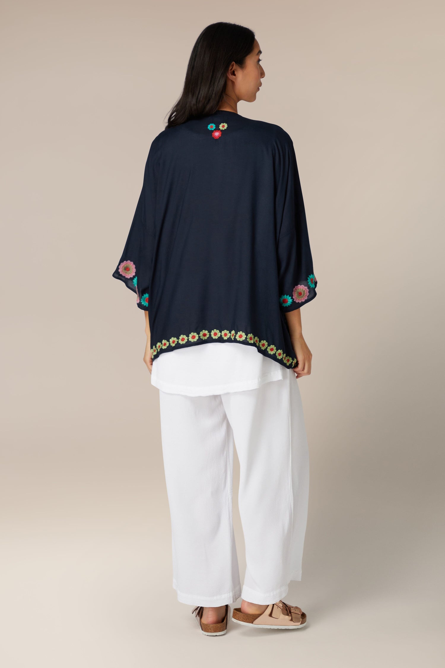 A woman standing with her back to the camera wearing a Lightweight Embroidered Flower Garden Kimono with colorful floral embroidery and white trousers.