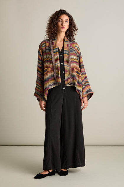 A person stands against a plain background, wearing a long black dress with buttons and the Hand Stitched Check Jacket, which features bold stripes, an open front, and wide sleeves, showcasing timeless style and artisanal craftsmanship.