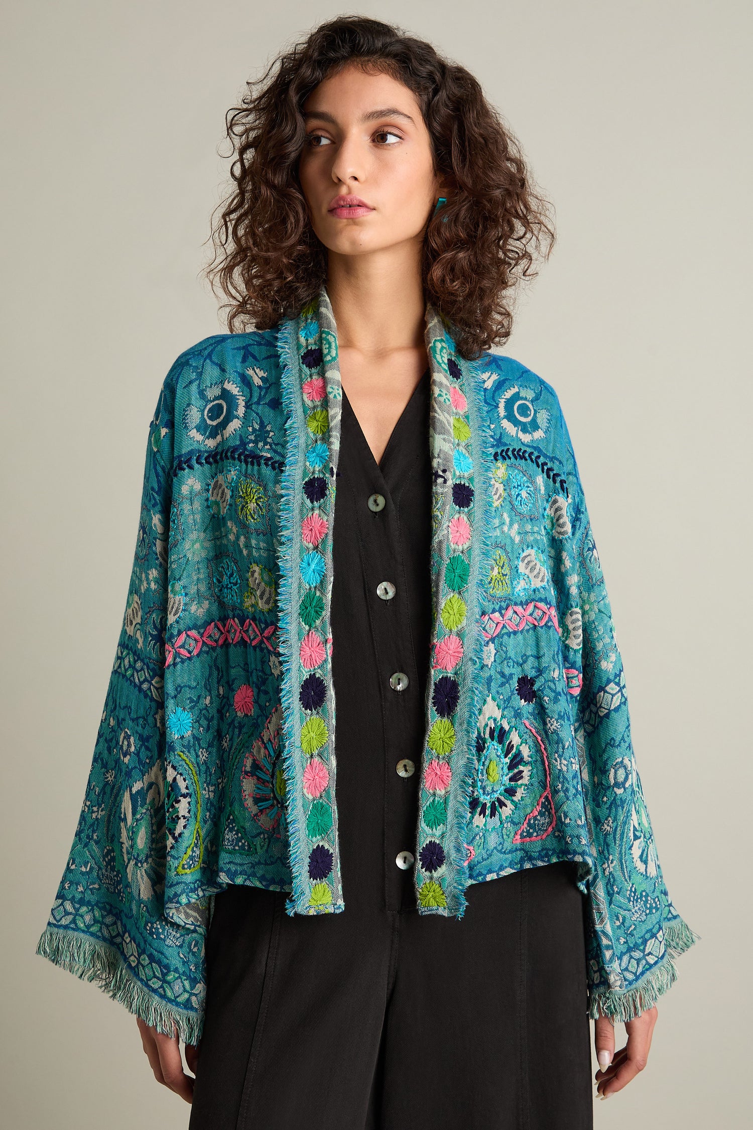 A person with curly hair wears a Cerulean Hand Embroidered Jacket over a black shirt, looking to the side against a plain backdrop.