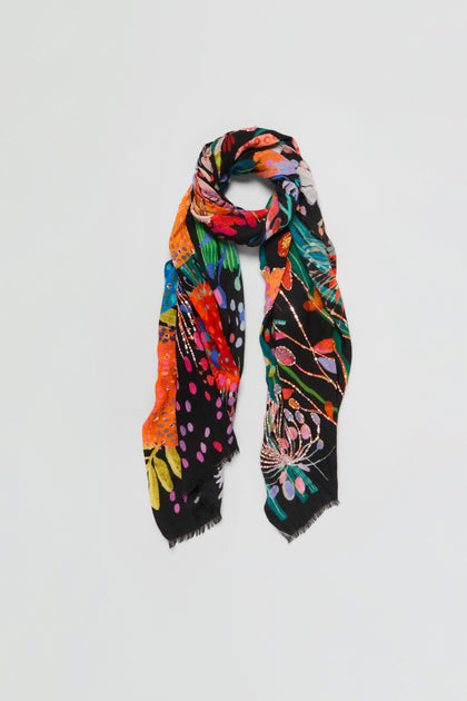 The Hand Embellished Wild Garden Wool Scarf, featuring a black base with a multicolored floral and abstract pattern and finished with frayed edges, is loosely draped and displayed against a plain white background.