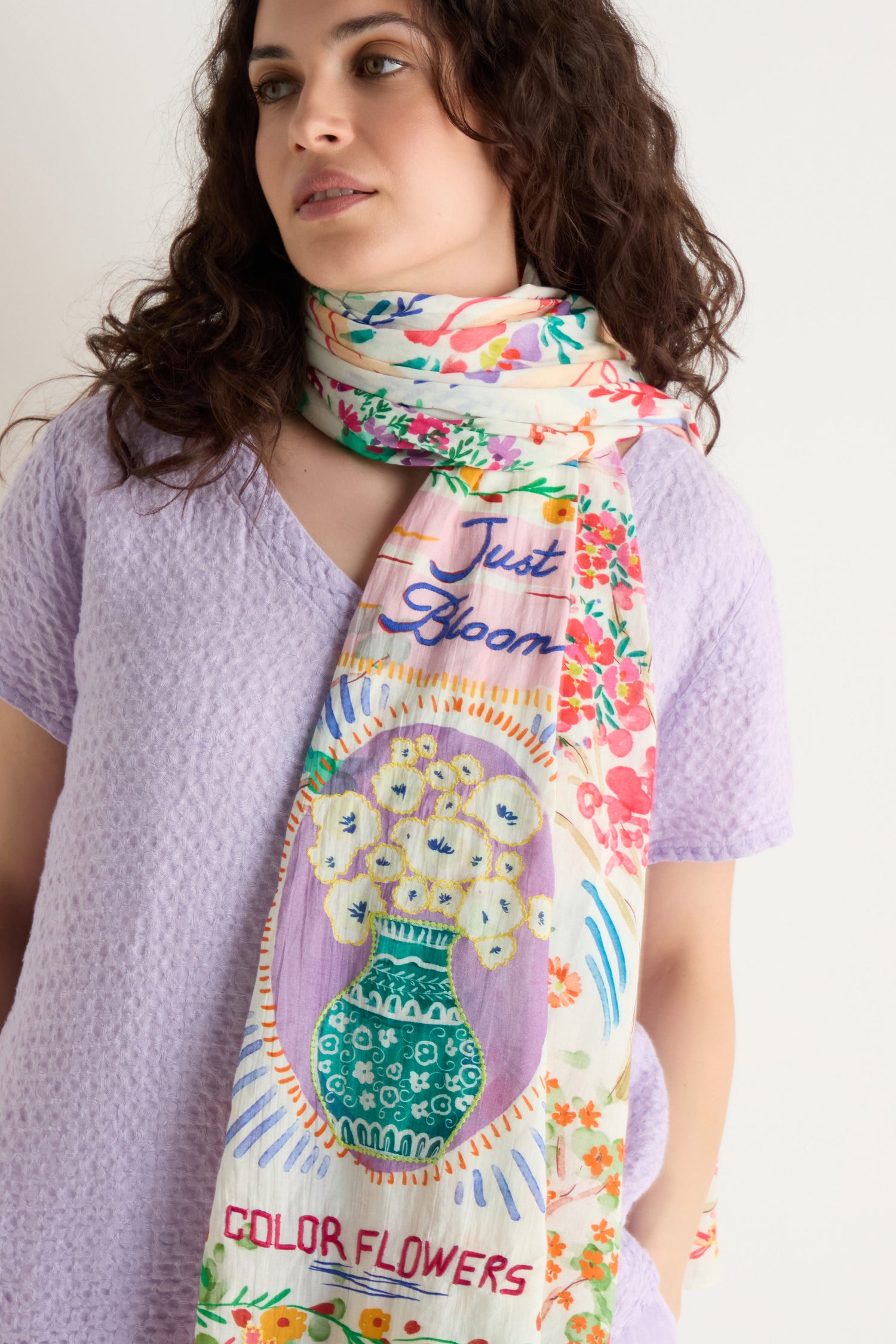 A woman wears a light purple textured top and the Floral Bouquet Cotton Scarf, featuring a vibrant floral print, vase illustration, and the words "Just Bloom" and "Color Flowers.