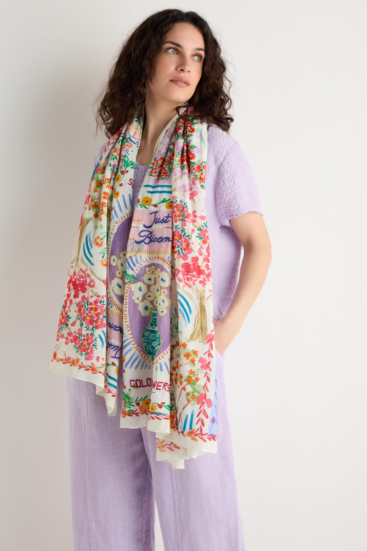 A woman stands against a plain white background, looking slightly to the side, wearing a light purple outfit and the Floral Bouquet Cotton Scarf featuring a colorful floral print.