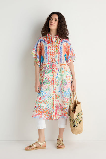 A woman stands against a white wall wearing the Floral Bouquet Cotton Shirt Dress over white pants, paired with sandals and holding a straw bag.