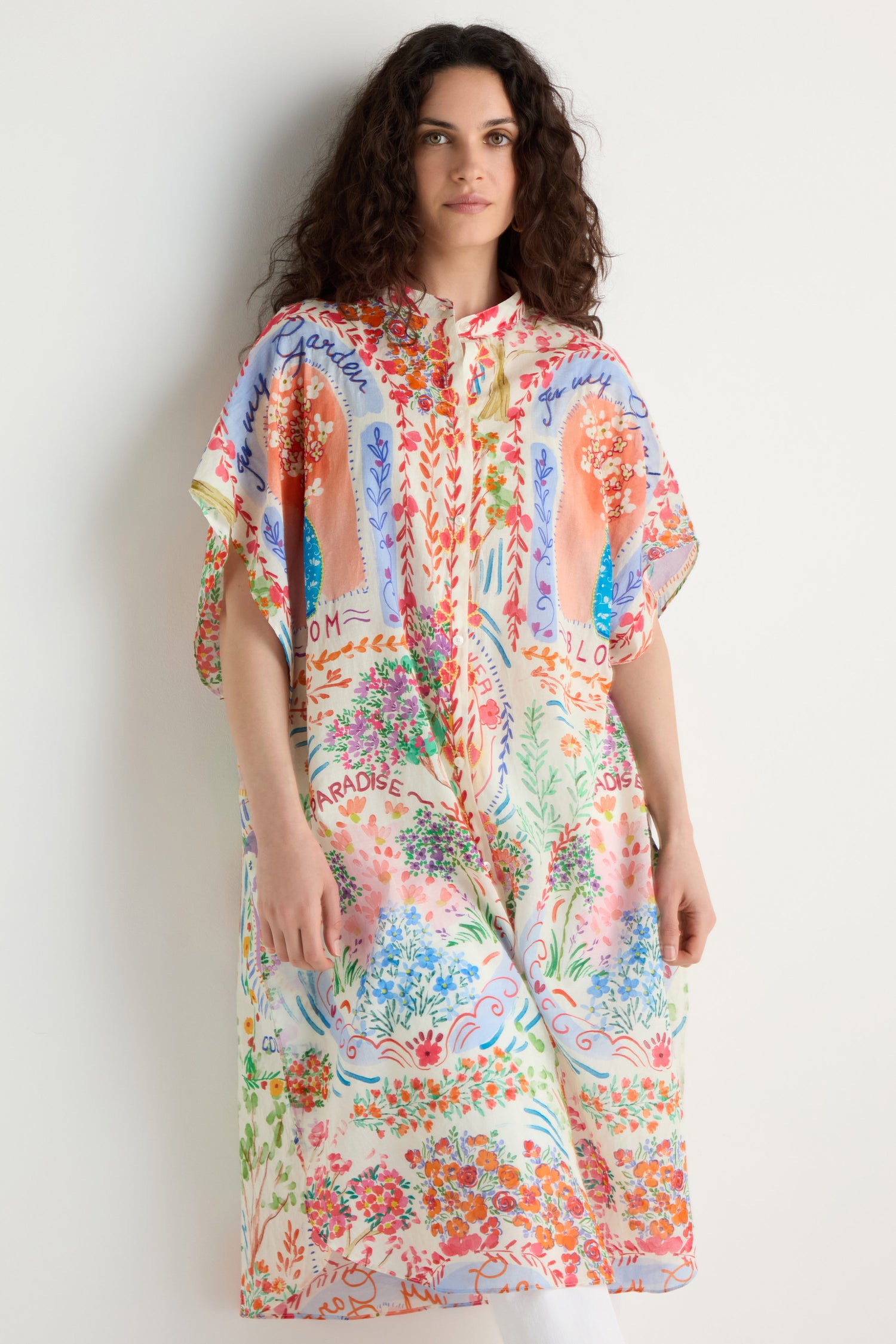 A woman with curly hair wears the Floral Bouquet Cotton Shirt Dress, featuring a colorful, loose fit and floral print, standing against a plain white background.