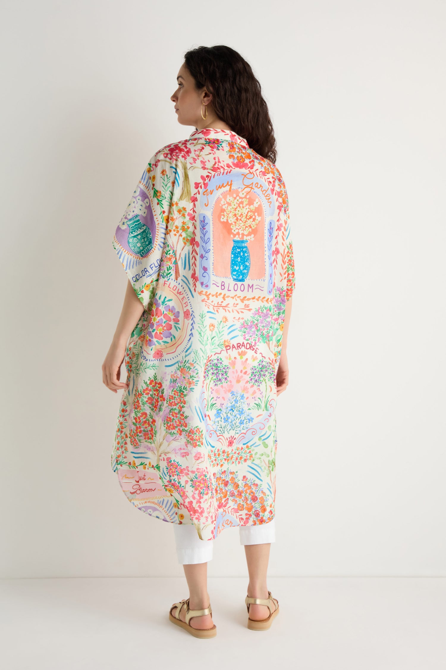 A woman stands facing away, wearing the Floral Bouquet Cotton Shirt Dress with a vibrant floral print over white pants and sandals, against a plain white background.
