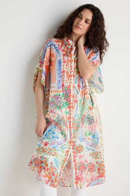 A woman stands against a white wall wearing the Floral Bouquet Cotton Shirt Dress, featuring colorful patterns and text designs, paired with white pants.