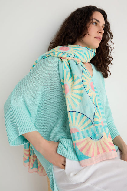 Woman with curly hair wears a light blue knit top, white pants, and the Embroidered Parasols Cotton Scarf featuring pastel hues, geometric patterns, and embroidery, standing against a plain white wall.