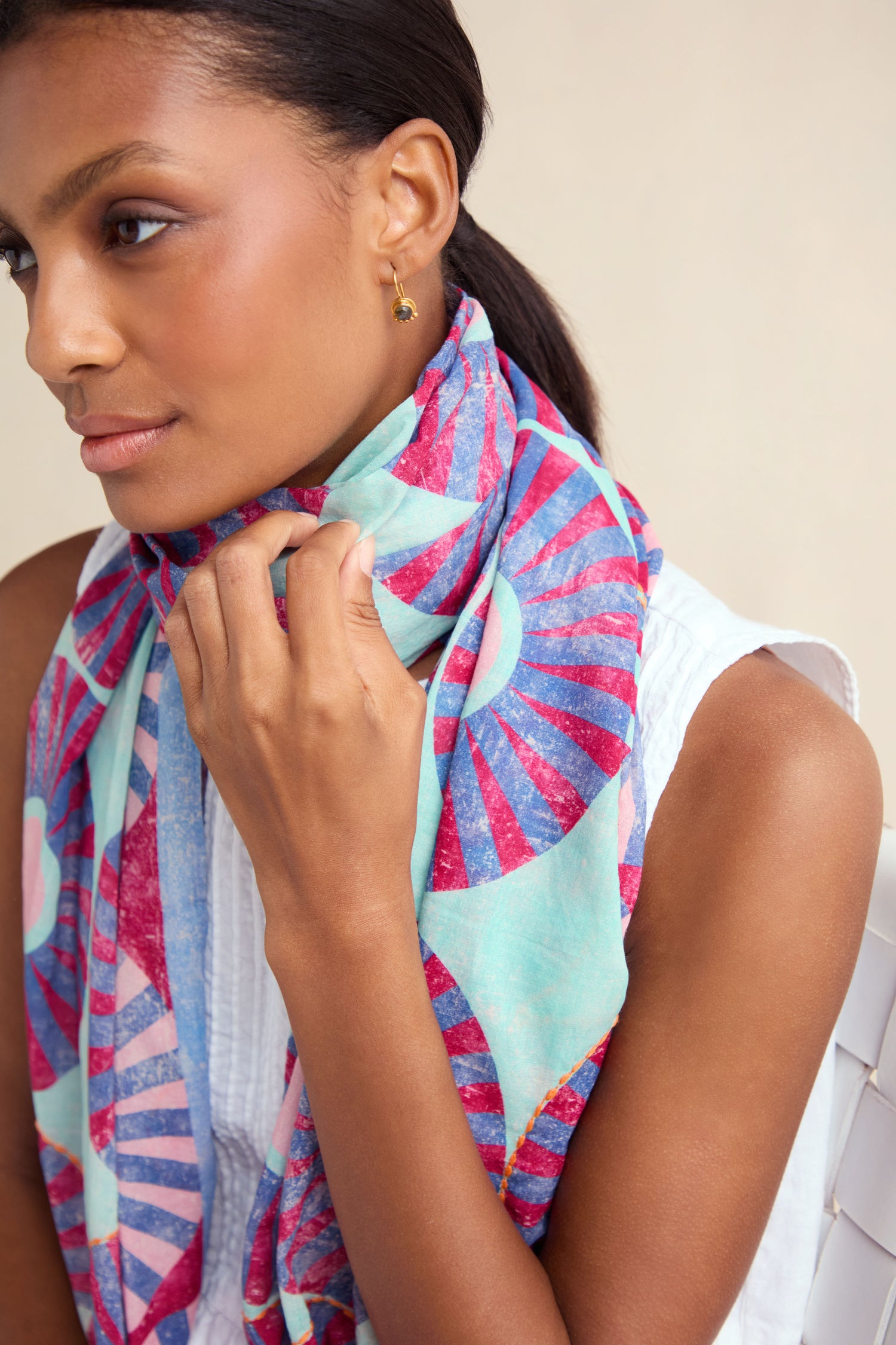A woman with dark hair tied back wears a sleeveless white top and the Embroidered Parasols Cotton Scarf, a colorful, lightweight cotton accessory draped gracefully as she rests her hand near her neck.