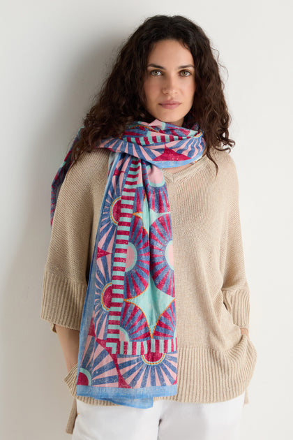 A woman with curly hair wears a beige sweater, white pants, and the Embroidered Parasols Cotton Scarf featuring geometric patterns, standing against a plain white wall.