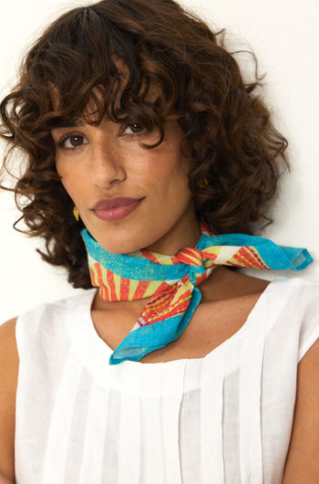 A woman with curly brown hair wears a white sleeveless top and the Embroidered Parasols Cotton Bandana, featuring a colorful blue and red print, tied around her neck as she stands against a plain background.