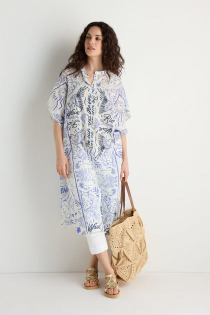 A woman wears the Paradiso Cotton Shirt Dress with embroidered details, standing against a white wall, styled with a woven tote bag and sandals.