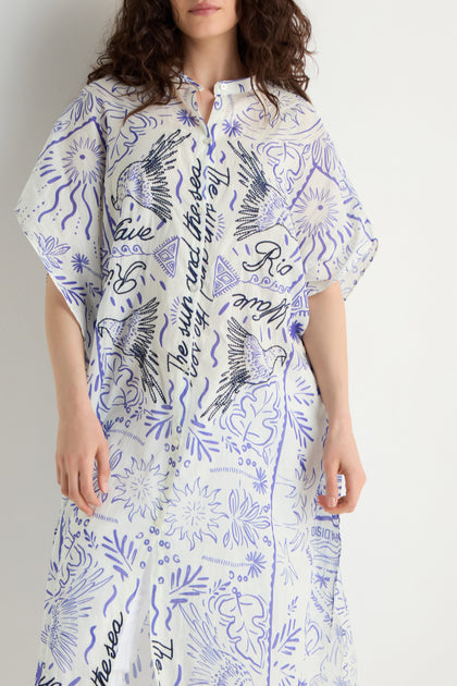 Someone stands against a white wall wearing the Paradiso Cotton Shirt Dress, featuring a relaxed silhouette, blue and black abstract designs with cursive writing, and embroidered detailing.
