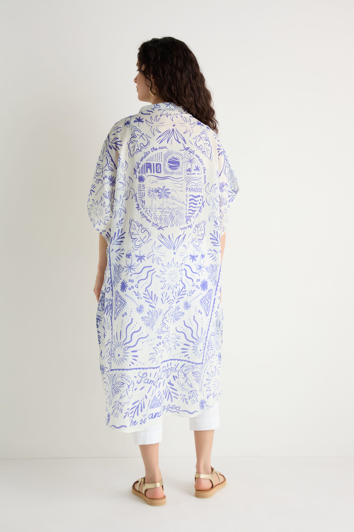 Person stands against a white wall, facing away, wearing the Paradiso Cotton Shirt Dress—a loose, knee-length cotton dress with blue graphic patterns and embroidery—paired with white pants and tan sandals.
