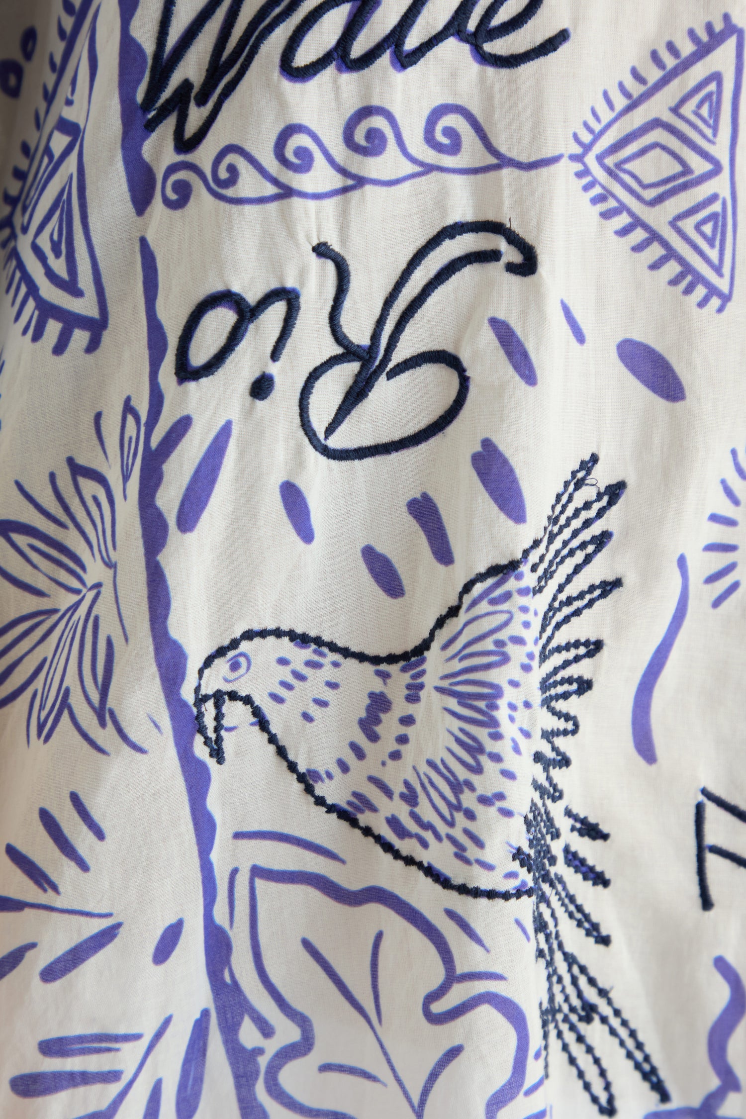 The Paradiso Cotton Shirt Dress is a white cotton dress with purple and black prints—bird, abstract shapes, partial text or symbols—and subtle embroidery for texture. It features a relaxed silhouette for an effortlessly stylish look.