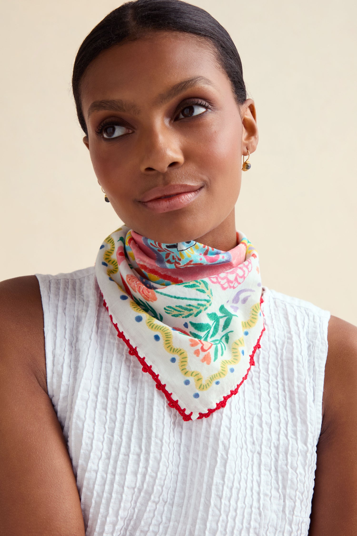 Woman wearing a white textured sleeveless top and the Embroidered Love In Bloom Cotton Bandana with red trim around her neck, looking slightly upward with a neutral expression.